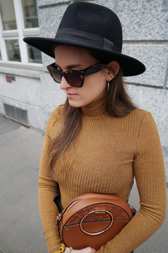 Andrea Steiner vom Mode Blog Strawberries 'n' Champagne aus der Schweiz trägt schwarze Lederhosen, einen braunen Rollkragen Pullover, dazu Tasche, Hut, Sonnenbrille und Plateauschuhe.